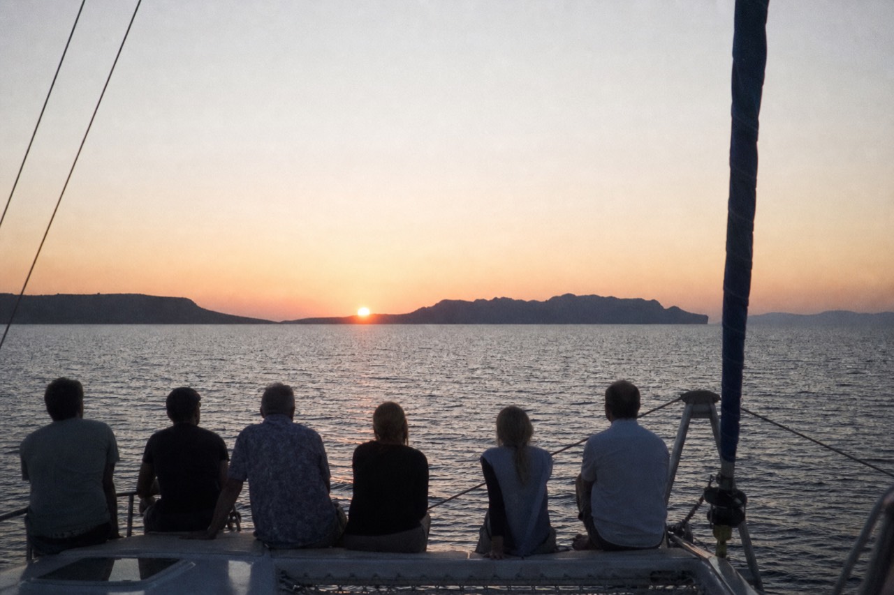 Silhouetted passengers along a catamaran rail at sea, backs to the camera, watching a low orange sun flatten onto the horizon beyond dark volcanic islands
