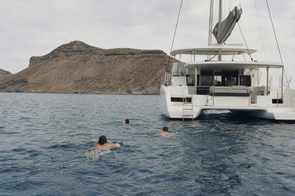 Swimmers in open water beside the lowered stern ladder of an anchored white catamaran, arms mid-stroke, a bare volcanic island rising behind them