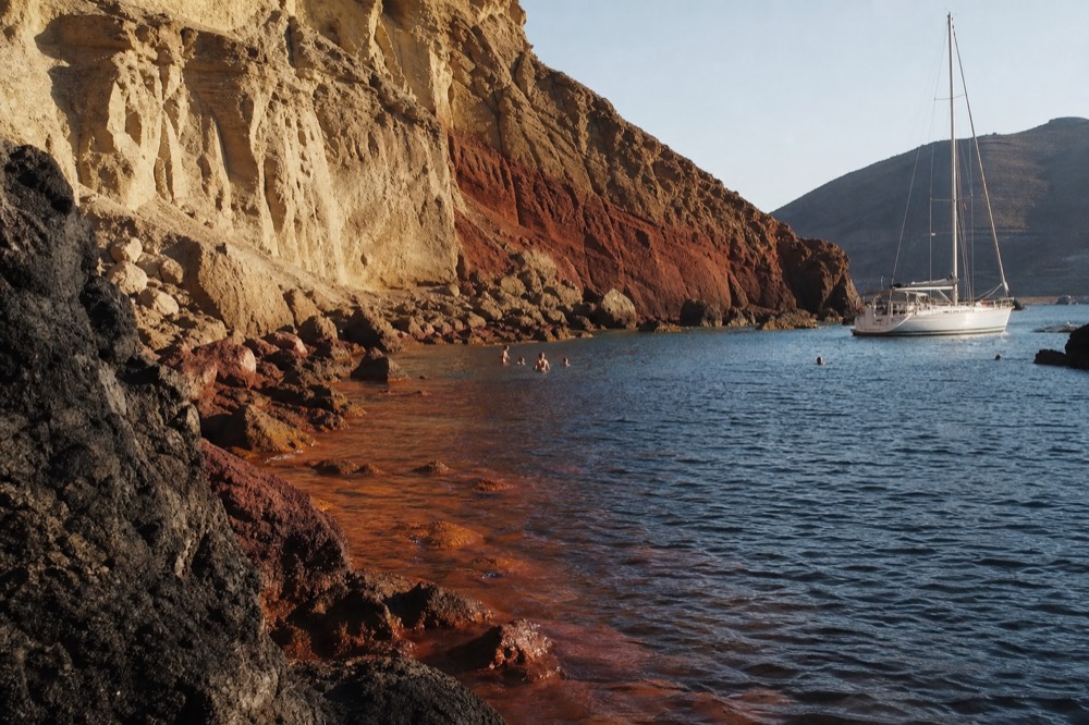 Rust-red mineral water fanning into deep blue sea inside a small volcanic cove, ochre-stained rock ledges rising steeply around a few distant swimmers