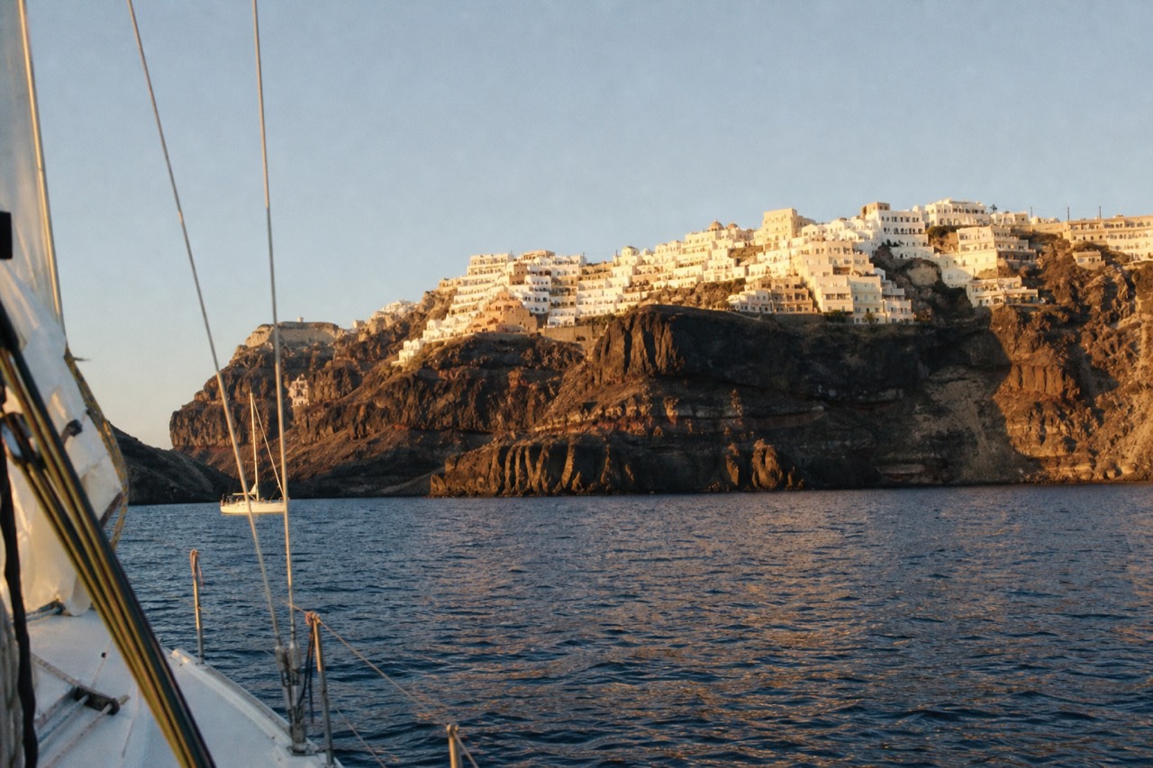 Whitewashed village stacked along a high cliff edge seen from sea level at golden hour, warm light on the terraces, shadowed rock falling to calm water