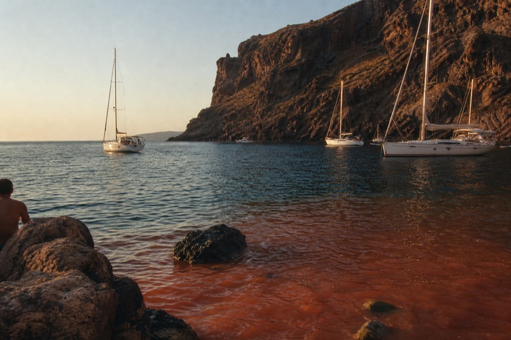 Rust-red mineral water meeting clear blue sea in a sheltered volcanic cove, stained ochre rock rising steeply behind the shallows