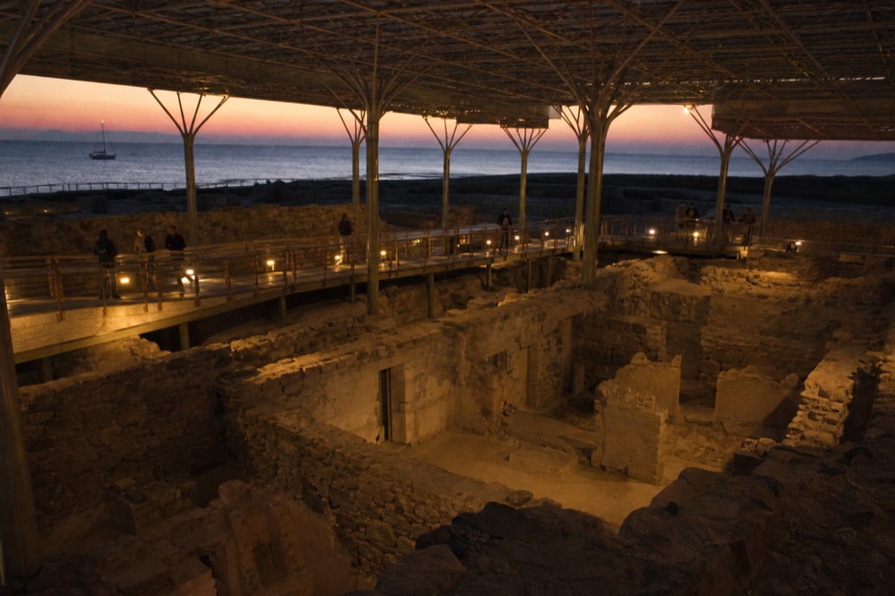 Excavated stone streets and two-storey walls of an ancient buried town beneath a wide modern shelter roof, low lamps lighting raised walkways above the ruins