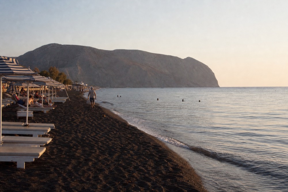 Rows of striped umbrellas on coarse black volcanic sand beside flat clear water, a low grey headland closing the far end of the bay