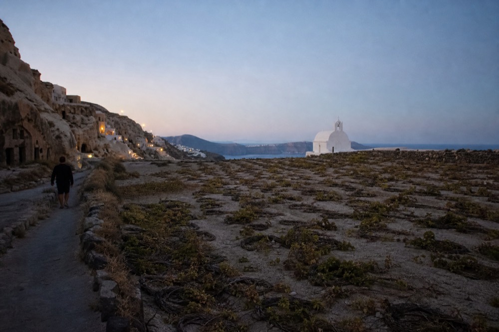Low basket-woven grapevines coiled flat on pale volcanic soil across an open field, a dry stone wall and a distant white chapel under hard midday sun
