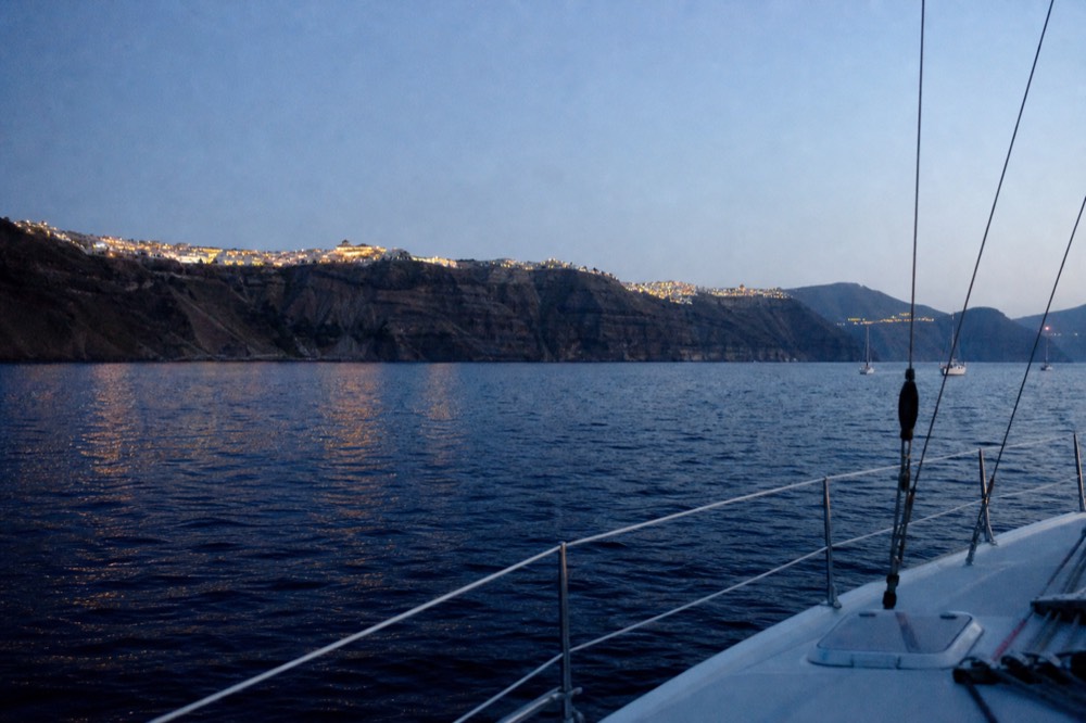 Deep blue twilight over a caldera rim, village lights strung along the clifftop and reflected in dark water below