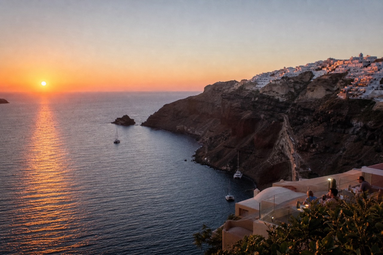 The sun as a flattened orange disk just above the sea, a whitewashed cliff village catching pink light along a high volcanic rim