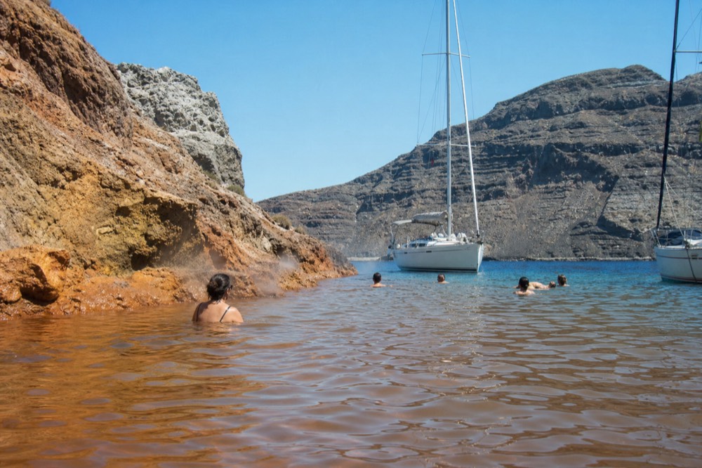 Swimmers treading warm rust-tinted water in a shallow volcanic cove, ochre rock steaming faintly at the waterline
