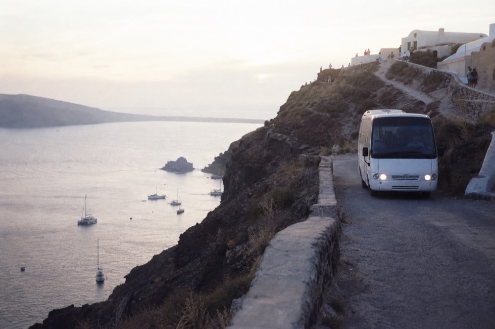 A white minibus waiting on a narrow cliff road above a red bay at dusk, headlights on, dark volcanic slope falling away to moored boats below