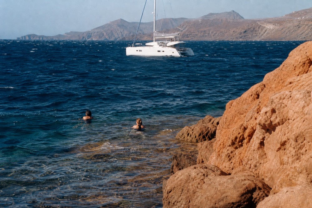Swimmers in dark suits treading rust-tinted shallow water below ochre volcanic rock, an anchored white catamaran fifty metres behind them