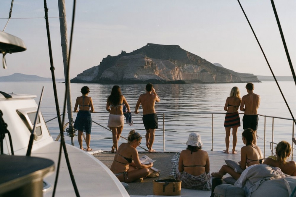 A busy stern deck on a catamaran at anchor, guests in swimwear leaning along the rail, a dark volcanic islet astern under hazy midday light