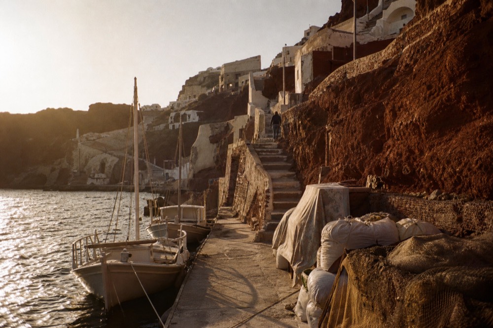 Red cliffs above a narrow concrete quay at a tiny fishing bay, wooden boats tied bow-in and steep switchback stairs climbing out of frame