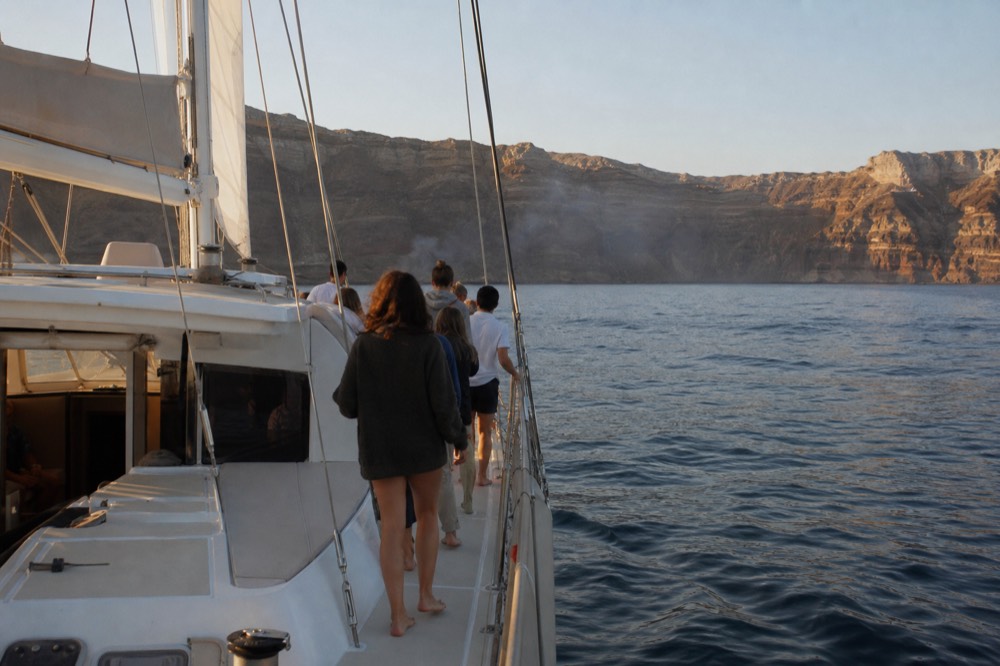 Guests queueing barefoot along a side deck toward a smoking stern grill at anchor, volcanic island low behind them in hazy late-afternoon light