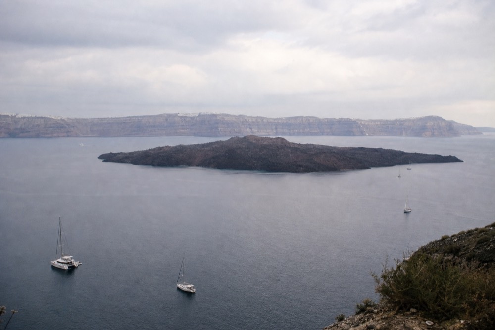 A low black volcanic island with a bare rocky summit sitting in the middle of calm water, pale caldera cliffs ringing the horizon behind it