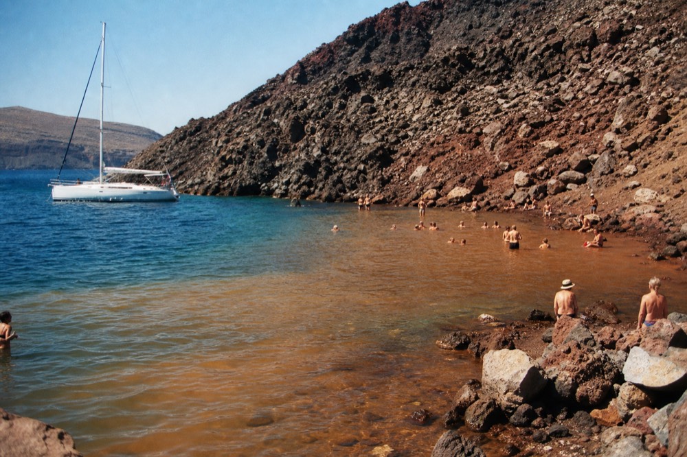 Rust-red mineral water meeting clear blue sea in the hot springs cove at Palea Kameni, ochre volcanic rock rising steeply above the shallow inlet