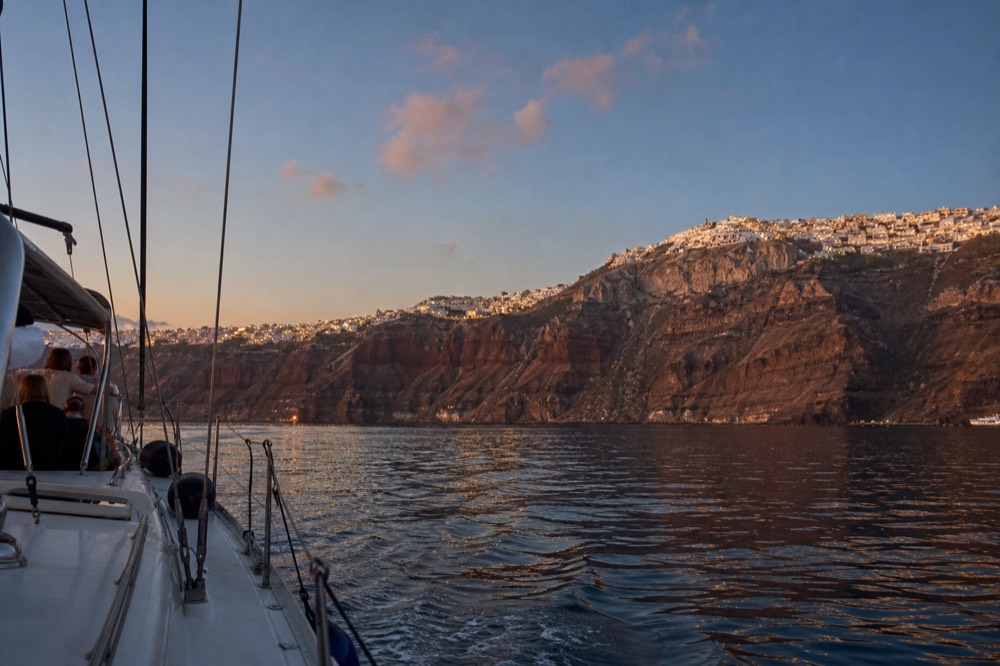 Warm village lights climbing the dark caldera rim during blue hour, seen low from the water with a faint wake trailing across still sea