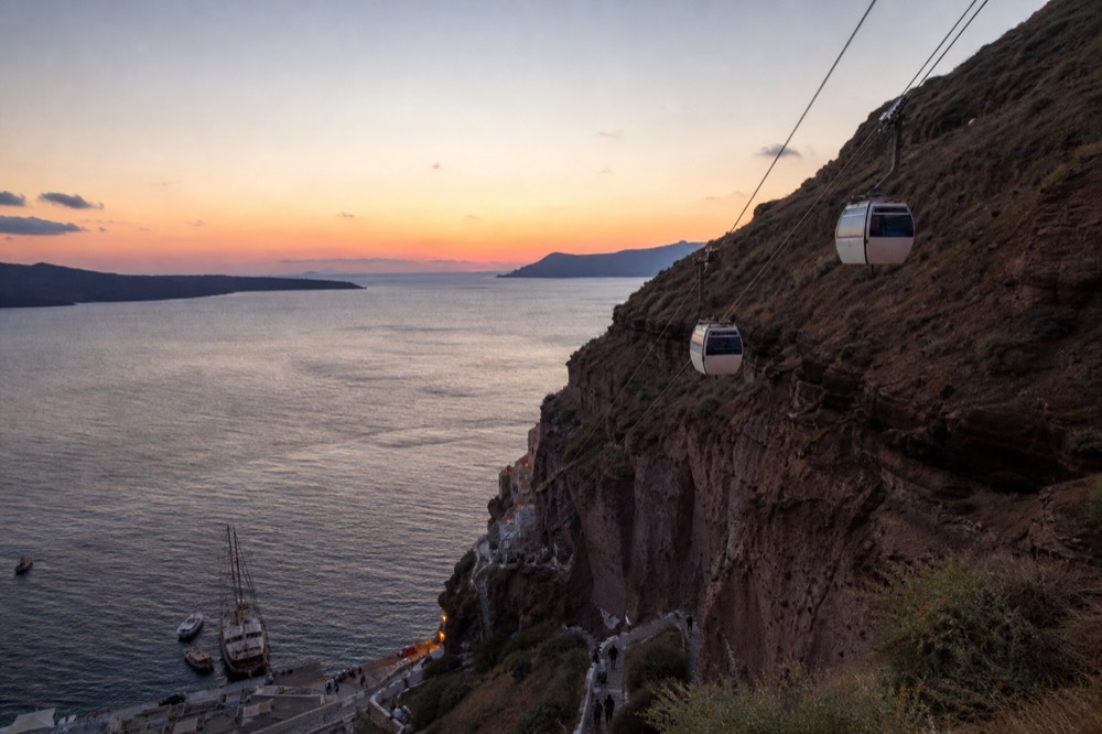 Cable car cabins climbing a sheer ochre cliff on twin cables, tiny figures on the switchback path far below them