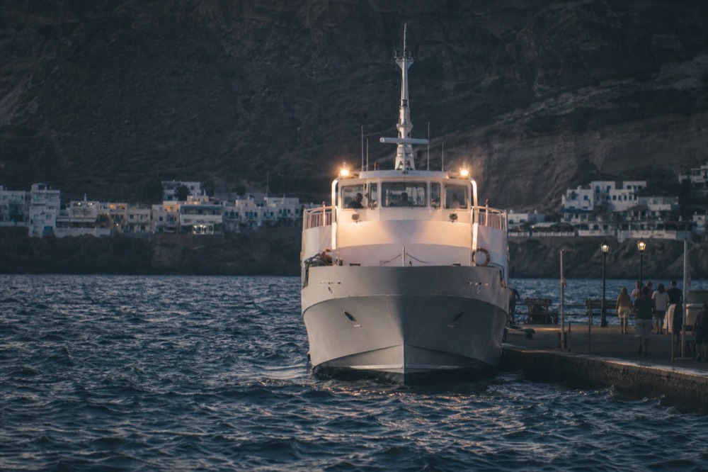 A ship's tender bow-on at a quay in fading light, deck lights on and the cliff above already deep in shadow