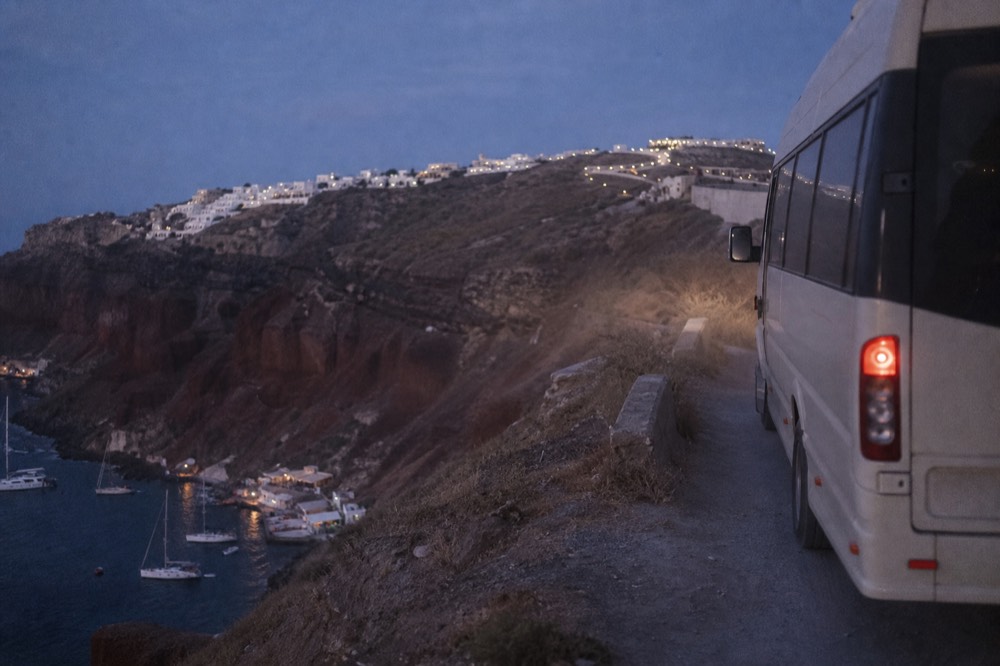 A minibus waiting on a narrow switchback road above a small red-cliffed bay, dust in the air and sailboat masts far below