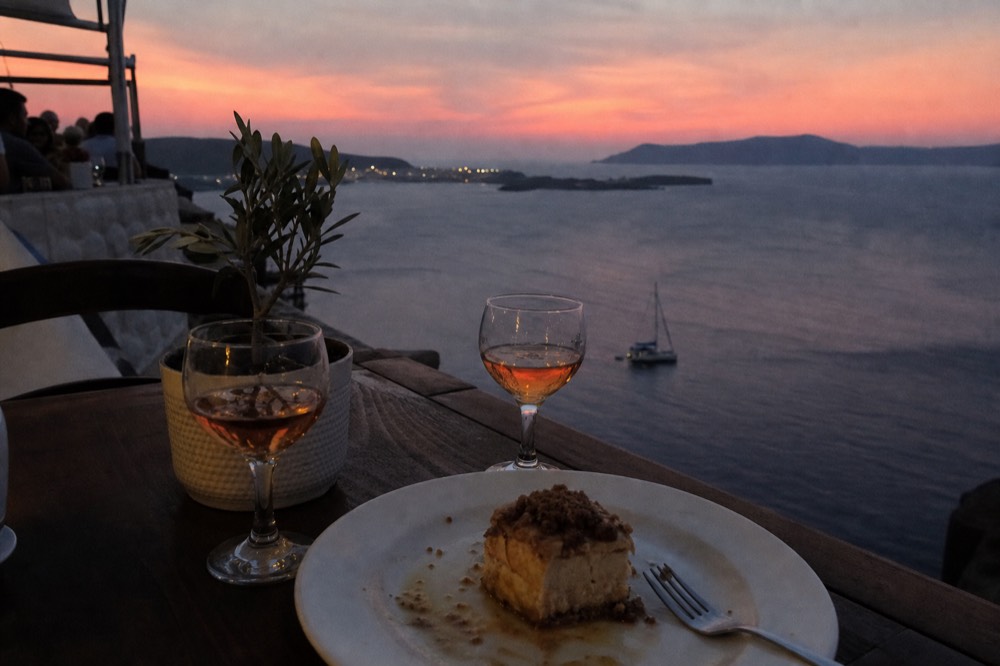 A dessert plate and two small glasses of amber wine on a deck table after dark, the caldera rim behind dotted with distant lights