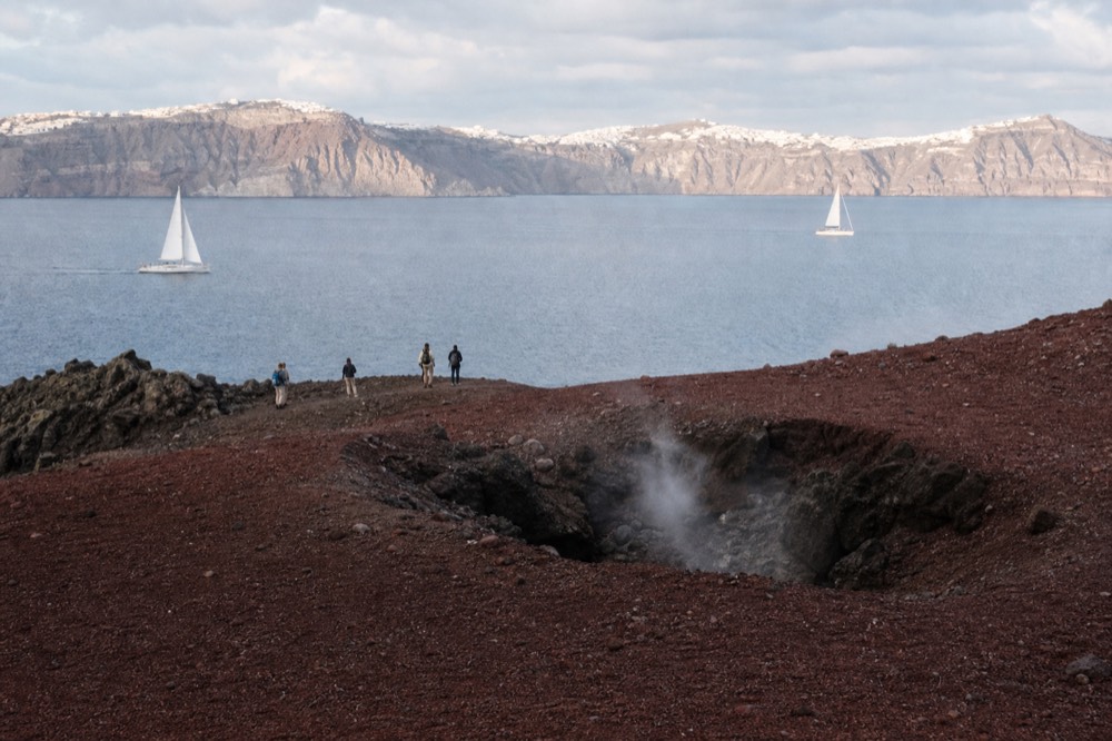 Faint steam drifting from a fissure in dark red volcanic gravel, sharp shadows and a hazy view of pale cliffs across the water beyond