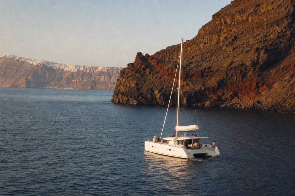 A white catamaran motoring slowly past the dark shoulder of a lava island in late afternoon, warm light raking across black rock and still water