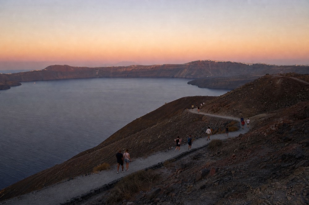 Walkers strung out along a pale gravel switchback on a bare volcanic cone, long shadows and the ringed caldera cliffs visible across flat water