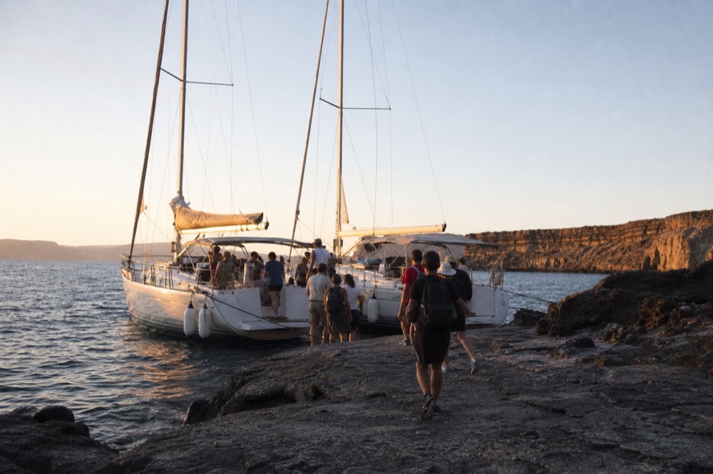 Two small excursion boats moored side by side against black lava rock, passengers stepping ashore onto a bare grey landing under a wide empty sky